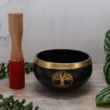 Black singing bowl with gold tree design and wooden mallet on a wooden surface.