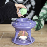 Purple oil burner with a lit candle inside, held by a hand wearing a black glove.