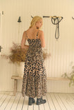 A woman stands with her back to the camera wearing a leopard-print maxi dress and black ankle boots in a rustic indoor setting with a white paneled wall, wooden bench, and potted plants.