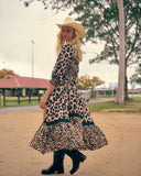 A woman wearing a leopard-print midi dress with a matching hat and black boots, standing outdoors on a dirt path.