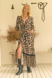 A woman modeling a leopard-print midi dress with a high slit, long sleeves, and a tie at the waist, paired with black boots, standing in front of a rustic cream-colored wall with western decor.