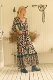 A woman modeling a long leopard-print maxi dress with a tie waist and black ruffled trim, standing in a rustic indoor setting with a bench and dried plants.