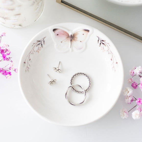 Jewelry dish with butterfly design holding rings and earrings on a light background with flowers.