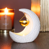 White ceramic moon-shaped lamp with a gold pendant on a wooden surface, candle in the background.