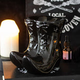 Black ceramic boot-shaped vase with star patterns on a dark surface with a candle and bottle in the background.