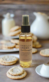 Vanilla creme body spray bottle on a table with cookies and a teapot in the background