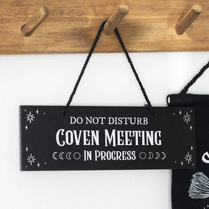 Black sign with white text 'Do Not Disturb Coven Meeting In Progress' hanging on a wooden peg rack.