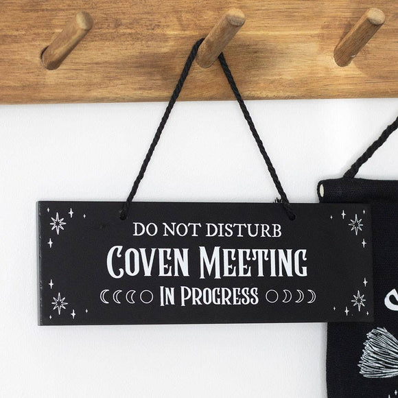 Black sign with white text 'Do Not Disturb Coven Meeting In Progress' hanging on a wooden peg rack.