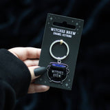 Keychain packaging with 'Witches Brew' design held by a hand against a dark background