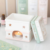 Decorative ceramic oil burner with a lit candle inside, on a light surface with books in the background.